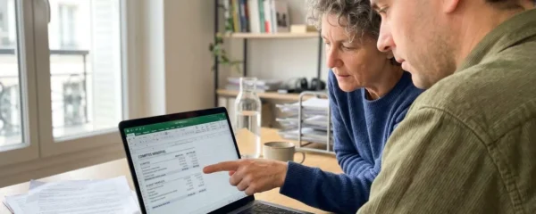 Vue par-dessus l'épaule de deux personnes consultant ensemble des documents financiers sur un ordinateur portable dans un bureau lumineux et contemporain