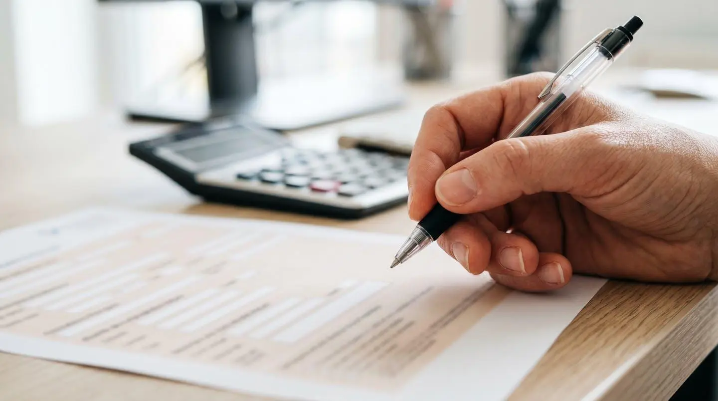 Gros plan sur une main tenant un stylo au-dessus d'un formulaire de demande de crédit avec texte complètement flouté et calculatrice moderne en arrière-plan